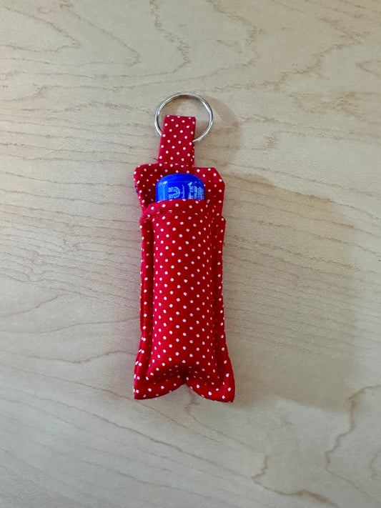 A small red fabric keychain pouch with white polka dots, holding a blue chapstick, resting on a light wood grain surface.